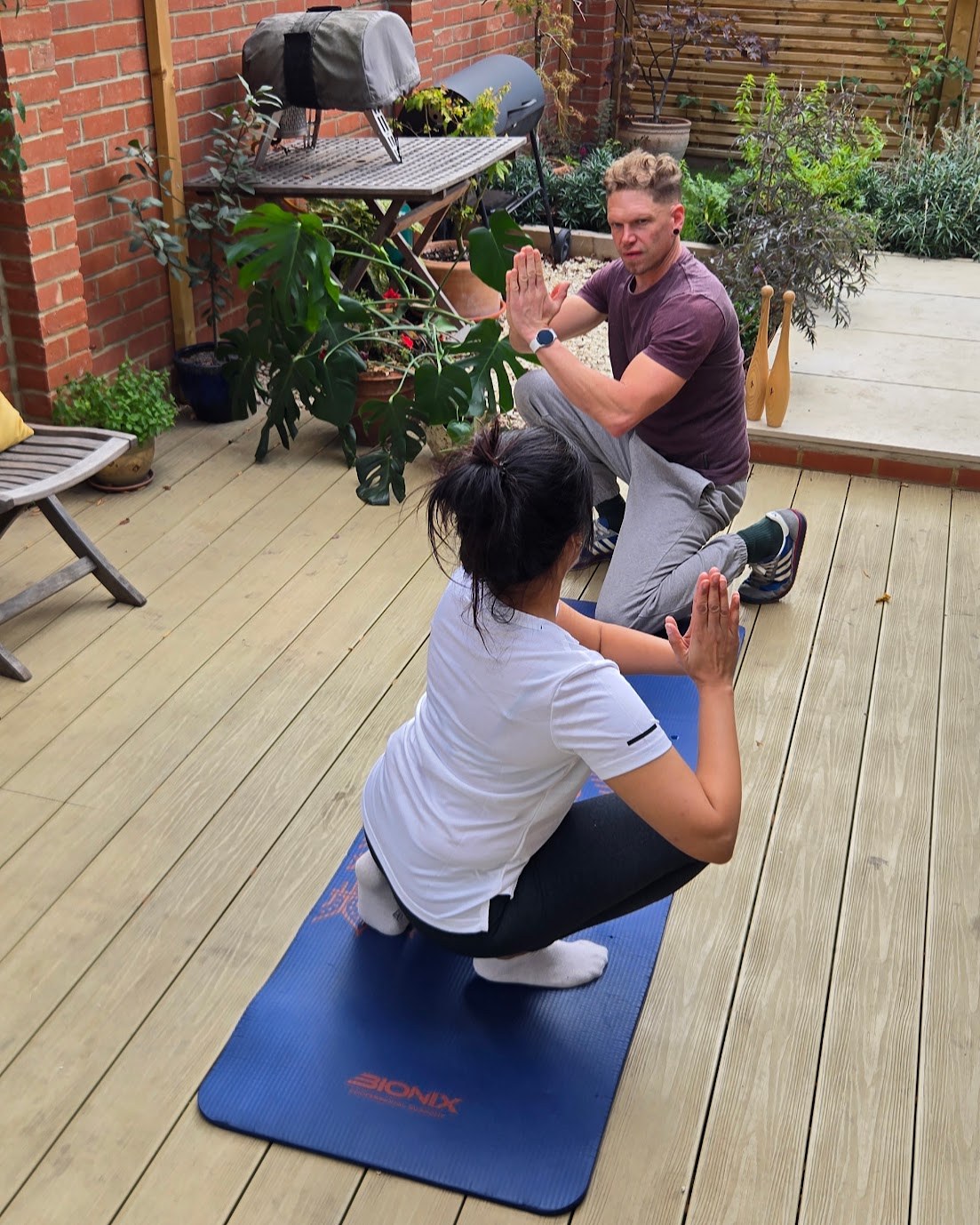 Matt coaching Dipti through a deep squat on a mat in the garden